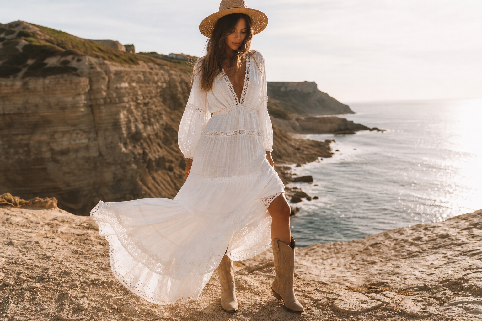 Femme en robe longue blanche, santiags beiges et chapeau de paille sur fond de côte ensoleillée